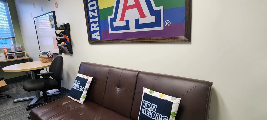 couch with two pillows, small circle desk with two roller chairs, and a UA rainbow flag above the couch