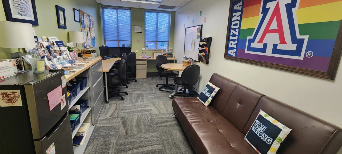 overview image of the HUB room, includes a couch with pillows, a fridge, flyers, desk space, and computers