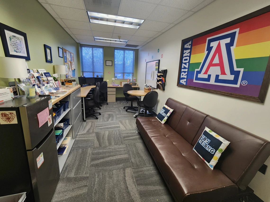 overview image of the HUB room, includes a couch with pillows, a fridge, flyers, desk space, and computers