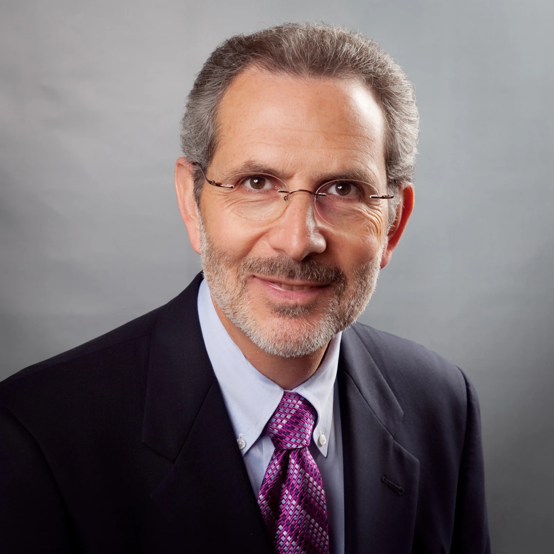 Headshot of man wearing suit with purple patterned tie and glasses smiling at the camera