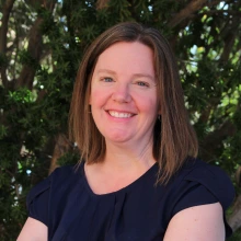 Image of woman with shoulder length hair standing in front of foliage wearing a blue shirt smiling at the camera.