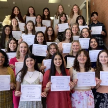 Image of about 25 college students standing on a stairway holding certificates in front of them and all smiling at the camera.