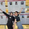 Two women arms out in front of poster